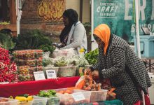 Duas mulheres organizam frutas e verduras em uma banca de feira ao ar livre.