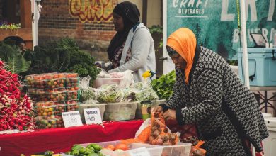 Duas mulheres organizam frutas e verduras em uma banca de feira ao ar livre.