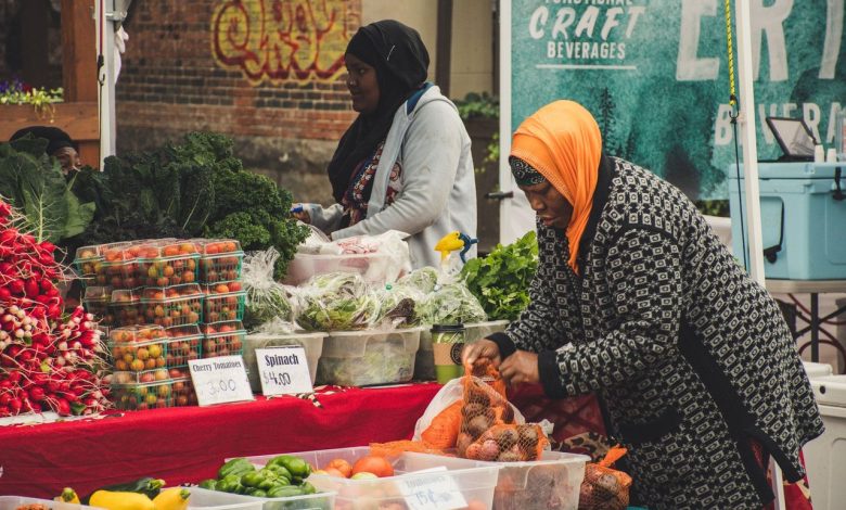 Duas mulheres organizam frutas e verduras em uma banca de feira ao ar livre.